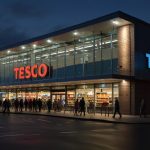 A brightly lit Tesco supermarket storefront at dusk, showcasing its modern design with prominent signage and welcoming entrance, bustling with shoppers.