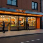 Modern Sainsbury’s convenience store with a brightly lit facade, showcasing fresh produce displays and eco-friendly signage in an urban UK setting.
