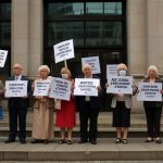 A crowd of low-income families and pensioners holding signs outside a DWP office, protesting the end of cost of living payments in 2025.