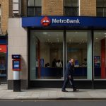 A modern Metro Bank branch with a brightly lit storefront, featuring large glass windows and the bank’s logo prominently displayed, located on a busy London street.