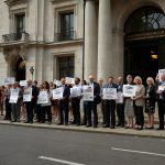 A diverse crowd of UK taxpayers holding signs and banners outside HM Treasury in London, advocating for a raise in the personal allowance for the 2025/26 tax year, with a focus on economic fairness.