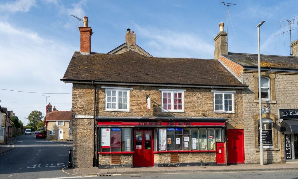 A row of stone-built shops and buildings on a high street in a rural English village, including a red-fronted storefront with windows, chimneys, and television aerials on the roofline.
