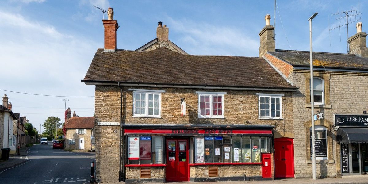 A row of stone-built shops and buildings on a high street in a rural English village, including a red-fronted storefront with windows, chimneys, and television aerials on the roofline.