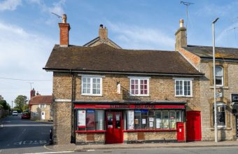 A row of stone-built shops and buildings on a high street in a rural English village, including a red-fronted storefront with windows, chimneys, and television aerials on the roofline.