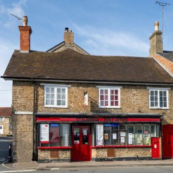 A row of stone-built shops and buildings on a high street in a rural English village, including a red-fronted storefront with windows, chimneys, and television aerials on the roofline.