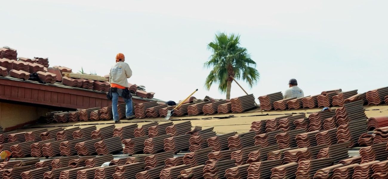 Two workers on a roof installing clay tiles, with a palm tree visible in the background.