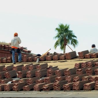 Two workers on a roof installing clay tiles, with a palm tree visible in the background.