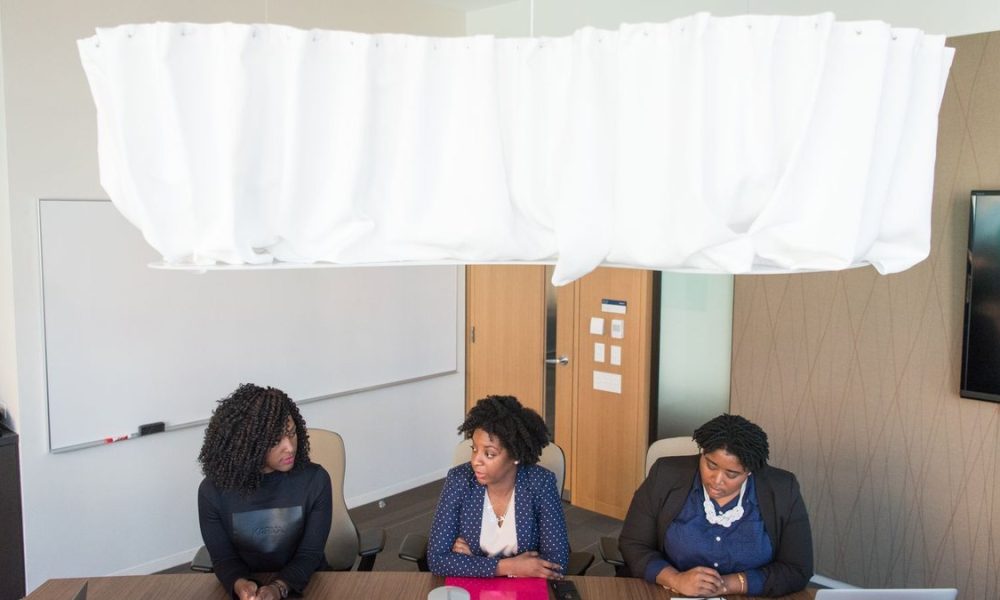 Three women in business attire sitting at a table in an office or classroom setting with a whiteboard and TV screen visible