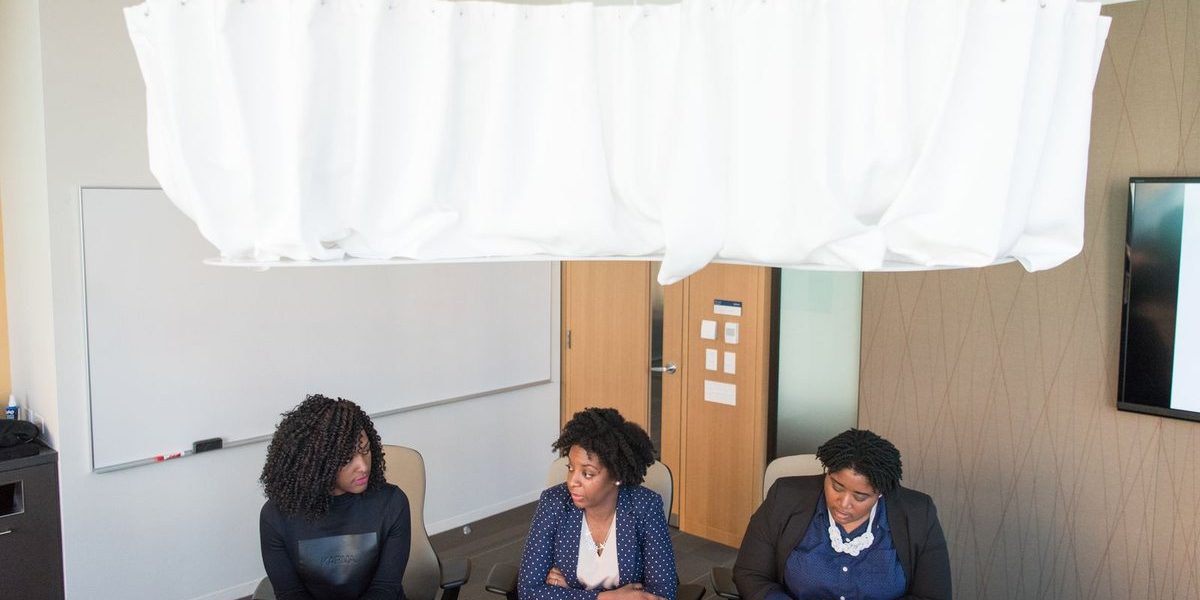 Three women in business attire sitting at a table in an office or classroom setting with a whiteboard and TV screen visible