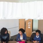 Three women in business attire sitting at a table in an office or classroom setting with a whiteboard and TV screen visible