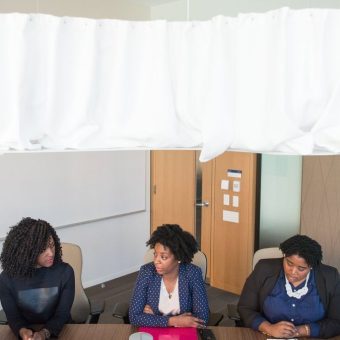Three women in business attire sitting at a table in an office or classroom setting with a whiteboard and TV screen visible